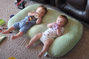 Two babies lying on a green pillow with toys around them.