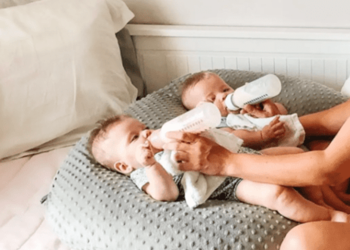 Two babies lying on a cushion with a person holding bottles