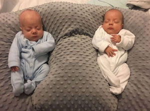 Two newborn babies in blue and white onesies lying on a textured gray pillow.