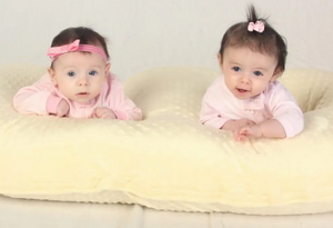Two babies lying on a yellow blanket wearing pink outfits and headbands.