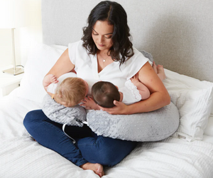 Woman breastfeeding two babies using a nursing pillow on a bed.