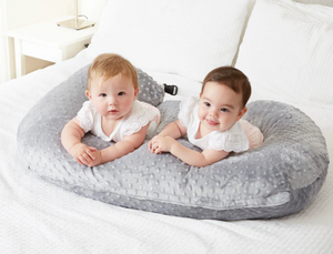 Two babies lying on a gray baby pillow on a white bed.