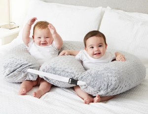 Two babies sitting on a gray body pillow on a bed.