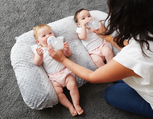 Two babies lying on a cushion with a woman holding a bottle and a pacifier.