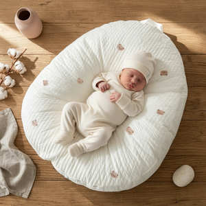 Newborn baby in a white outfit lying on a textured round pillow on a wooden floor.
