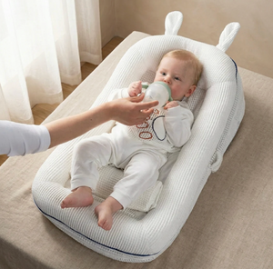 Baby in a white crib with a bottle, held by an adult's hand, in a softly lit room.