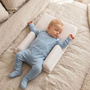A sleeping baby in a light blue footed sleeper lies on a white baby pillow with side supports on a beige linen bed.