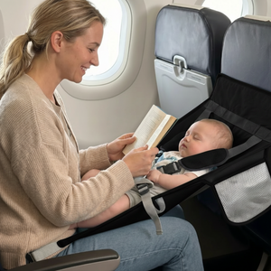 Woman reading a book to a baby in a portable bassinet on an airplane.