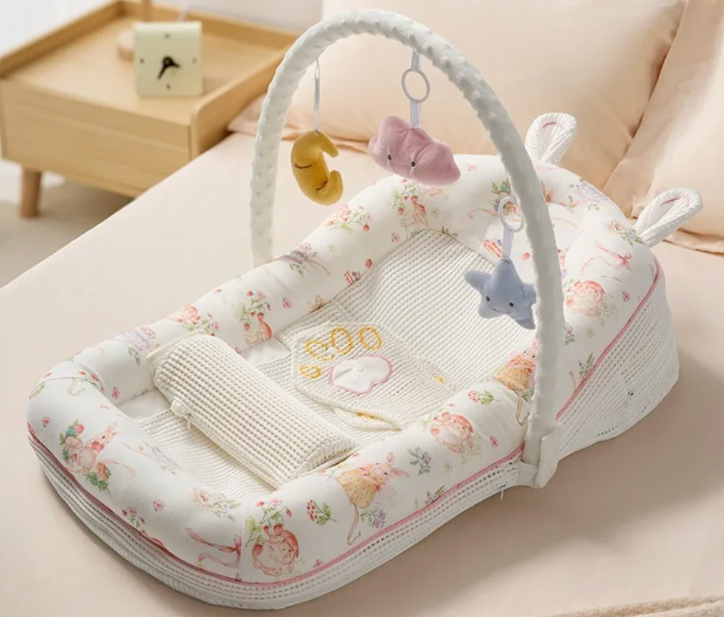A baby bassinet with an attached white arch activity toy, decorated with a soft pastel floral and animal print. The bassinet is styled on a beige bed with a wooden side table in the background.