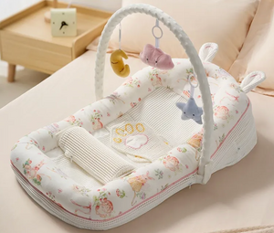 A baby bassinet with an attached white arch activity toy, decorated with a soft pastel floral and animal print. The bassinet is styled on a beige bed with a wooden side table in the background.