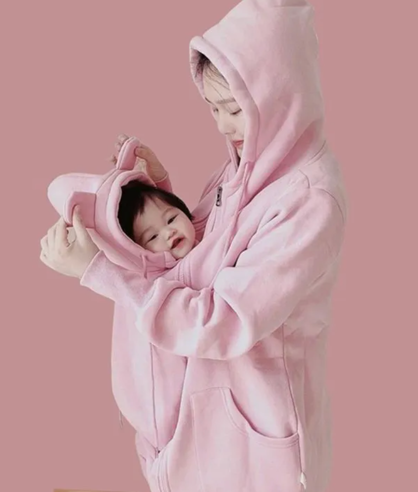 A woman wearing a pink zip-up hoodie with a baby carrier hood holds a baby in a matching pink hooded carrier, set against a solid pink background.