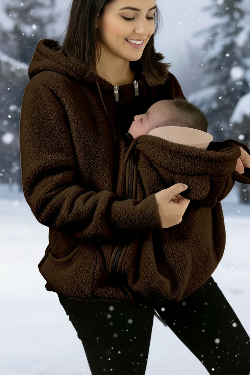 A woman wearing a dark brown sherpa fleece zip-up hoodie with a baby carrier pocket holding a sleeping baby, set against a snowy outdoor background with a flame graphic and '9.9' badge.