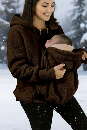 A woman wearing a dark brown sherpa fleece zip-up hoodie with a baby carrier pocket holding a sleeping baby, set against a snowy outdoor background with a flame graphic and '9.9' badge.