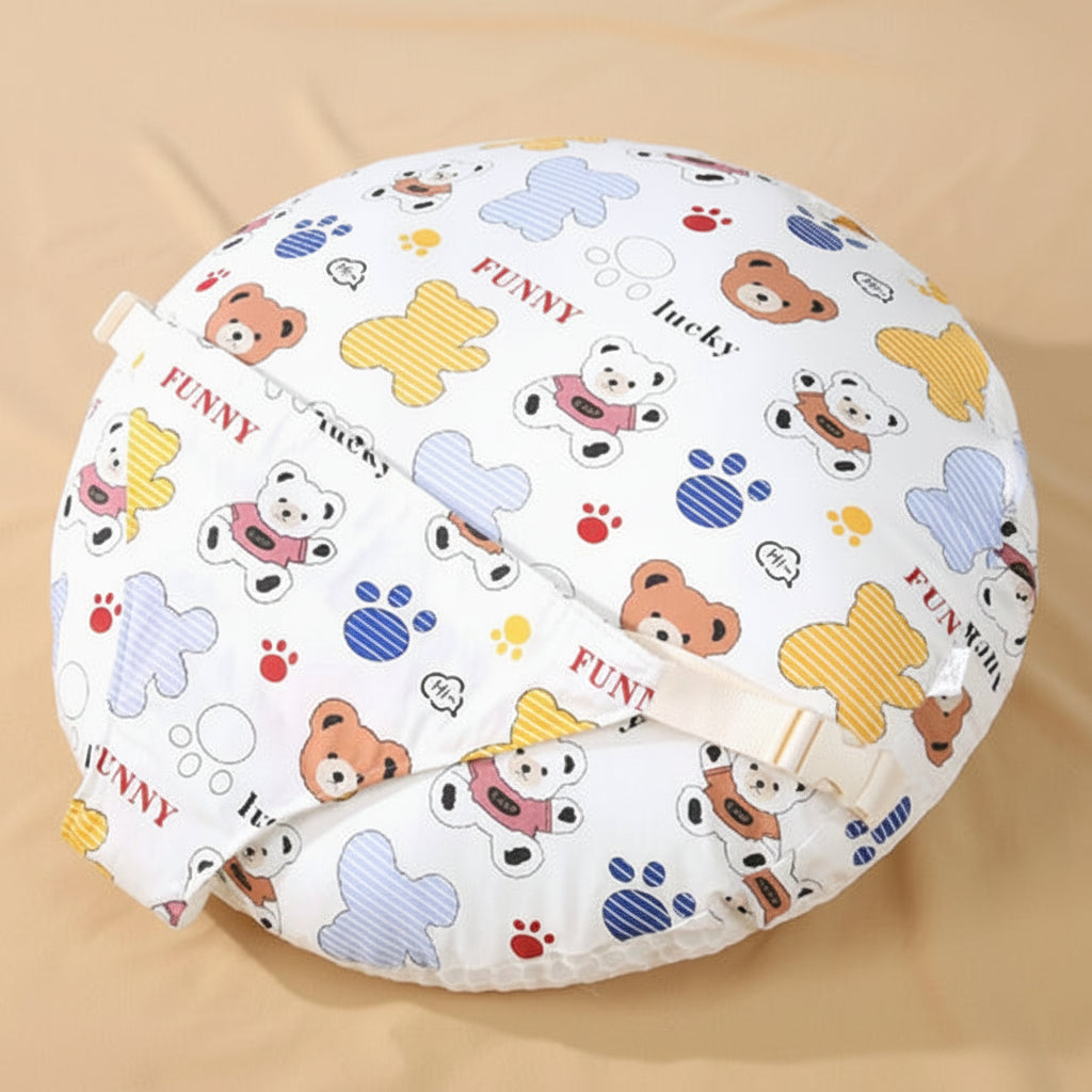 A round baby nursing pillow with a white background and a colorful cartoon bear print, featuring a beige strap with a plastic buckle. The image is set against a soft beige background 