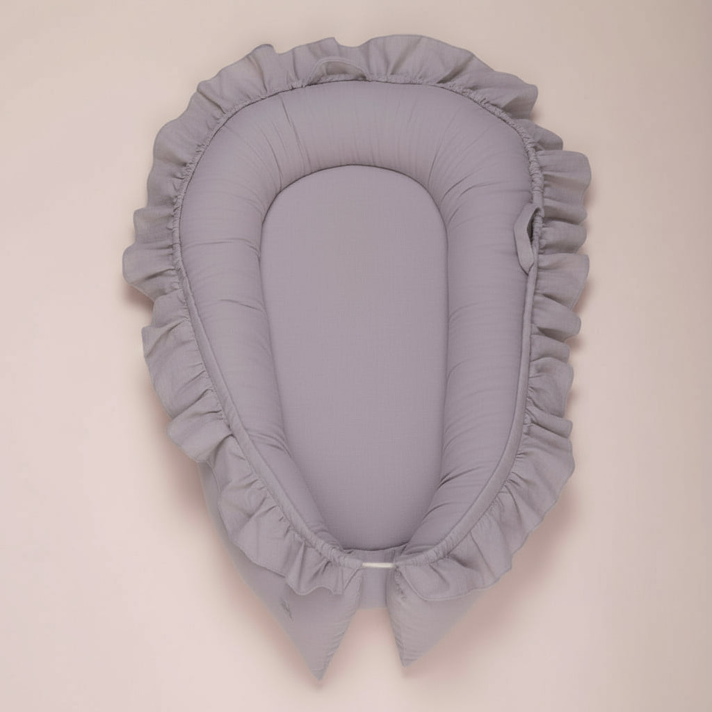 A newborn baby in a beige DreamCocoon baby lounger bed with decorative elements around, such as a green plant and a toy apple.