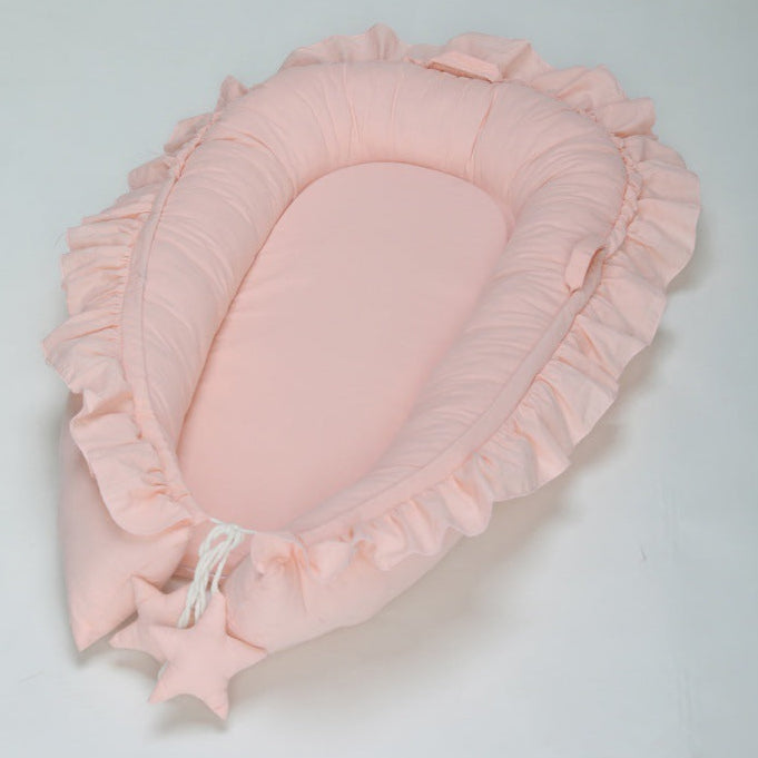 A newborn baby in a beige DreamCocoon baby lounger bed with decorative elements around, such as a green plant and a toy apple.