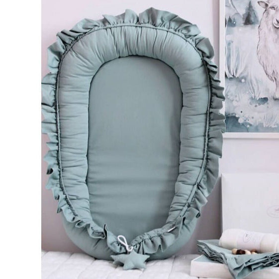 A newborn baby in a beige DreamCocoon baby lounger bed with decorative elements around, such as a green plant and a toy apple.