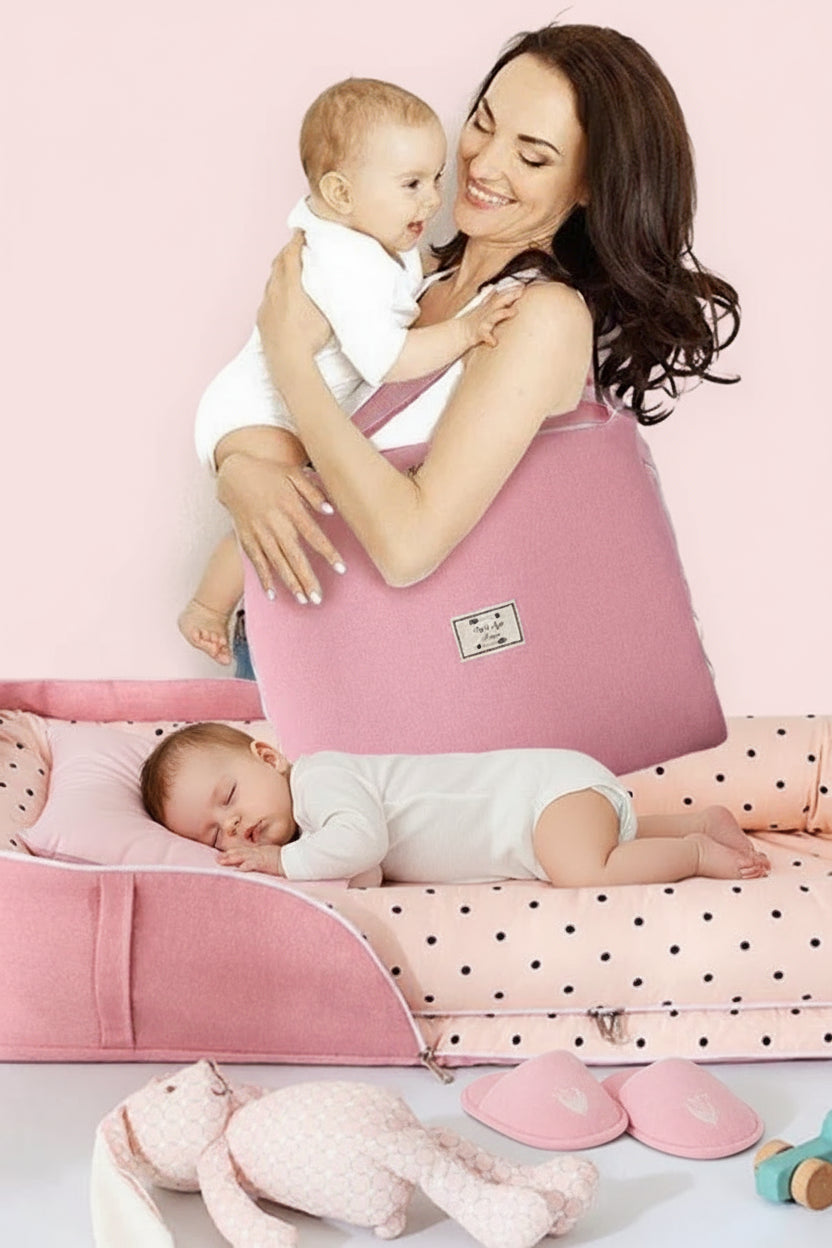 A baby travel sleep bed in pink color, with a woman holding a baby and sitting beside the bed, which appears to be made of cotton fabric. The bed is foldable and has a carry handle, indicating portability.